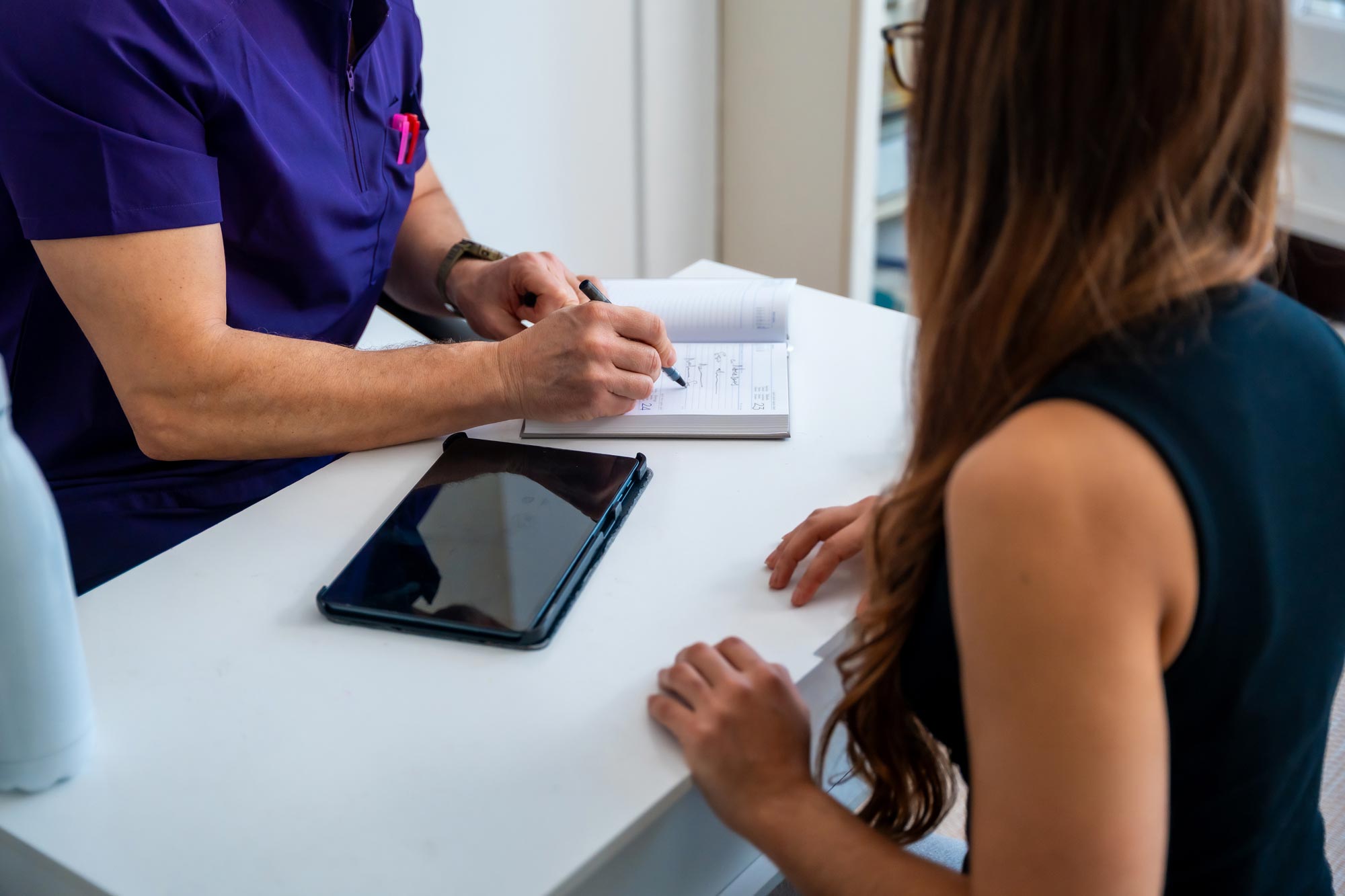 A consultant writing notes in a planner beside a tablet while a client looks on, going through next steps together