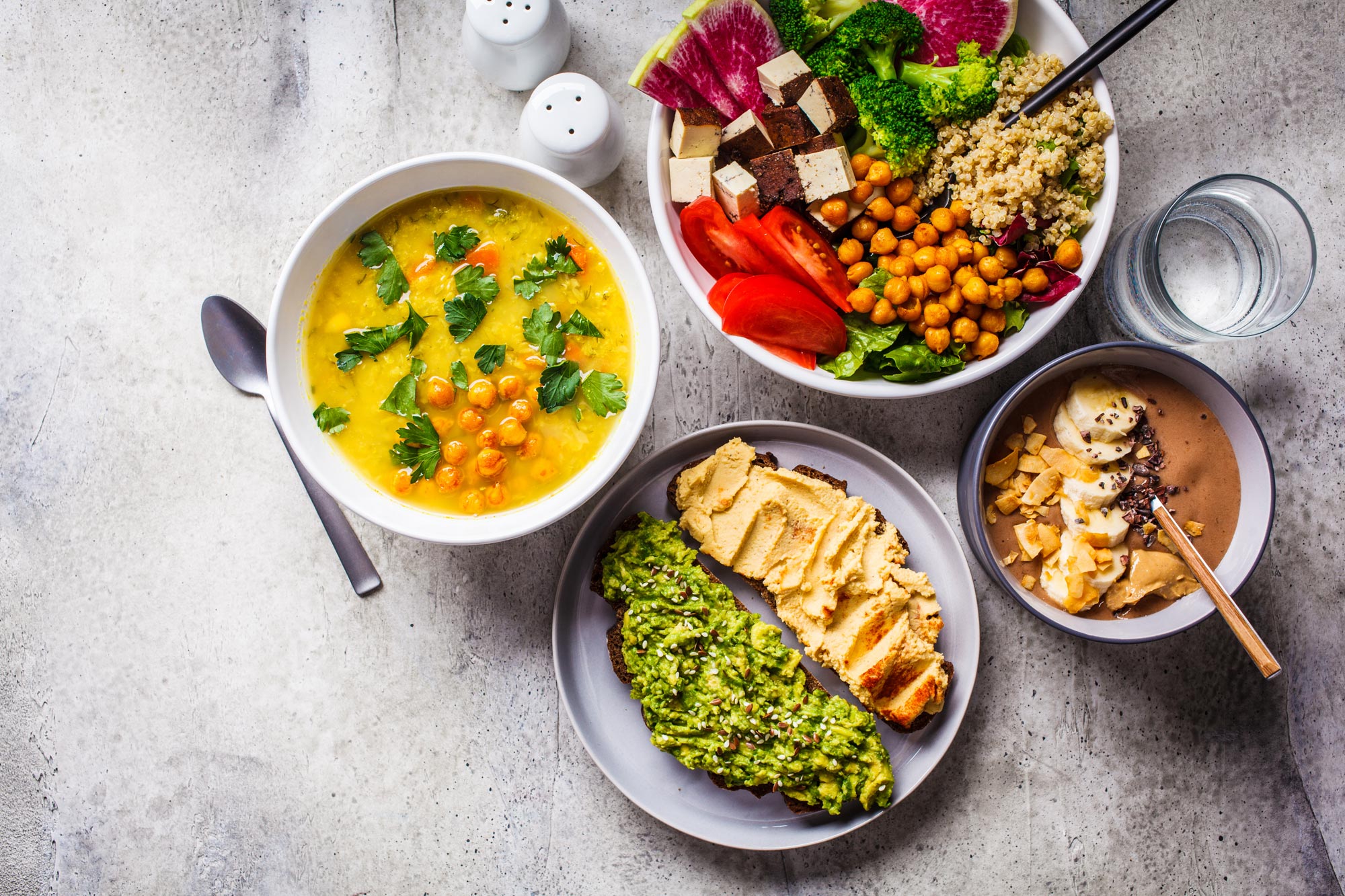 A table of plant-based dishes: lentil soup, a tofu and chickpea bowl, avocado and hummus toast, and a smoothie bowl