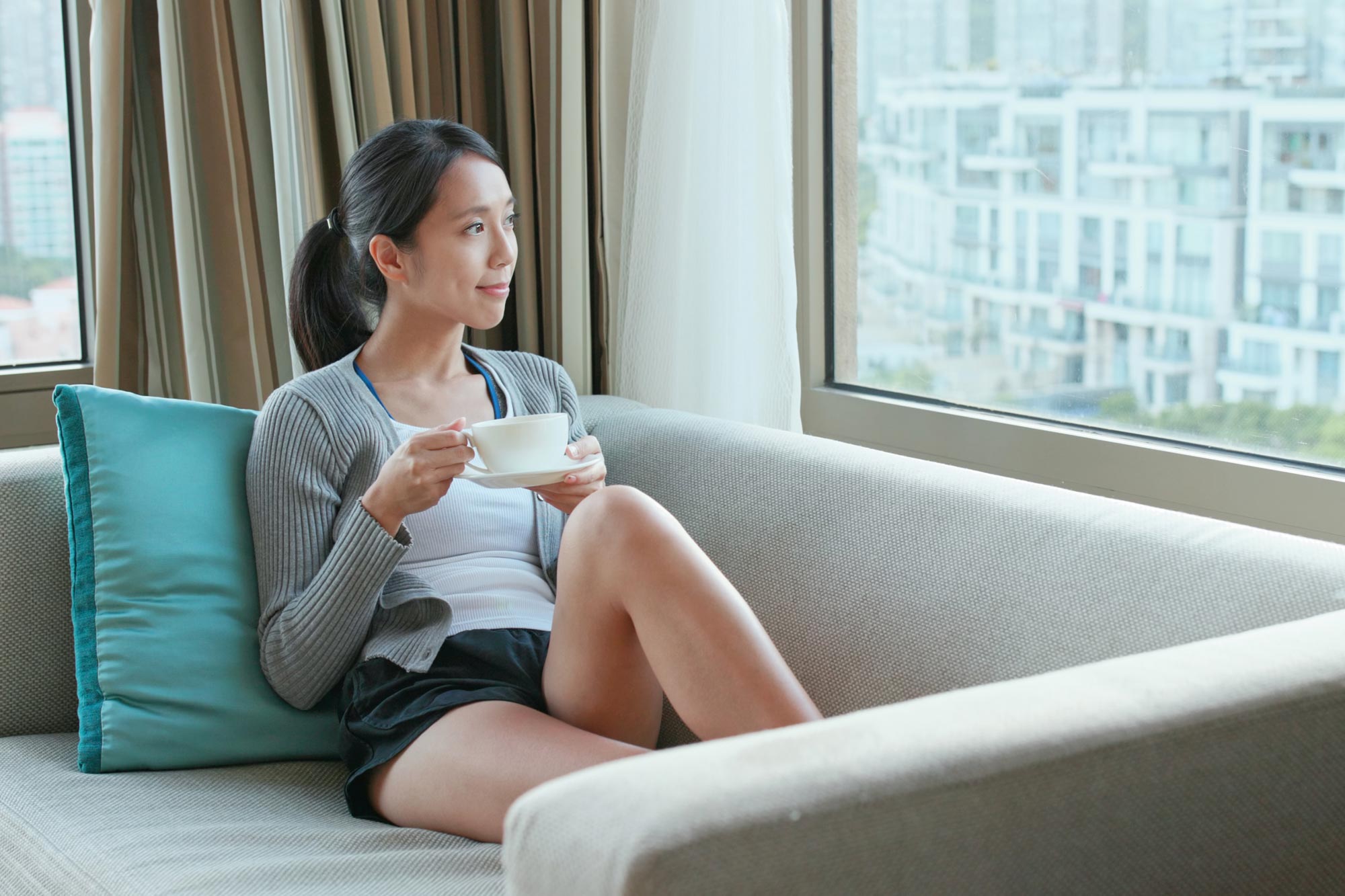 A woman relaxes on a sofa with a cup of tea, looking out of a window over city apartments