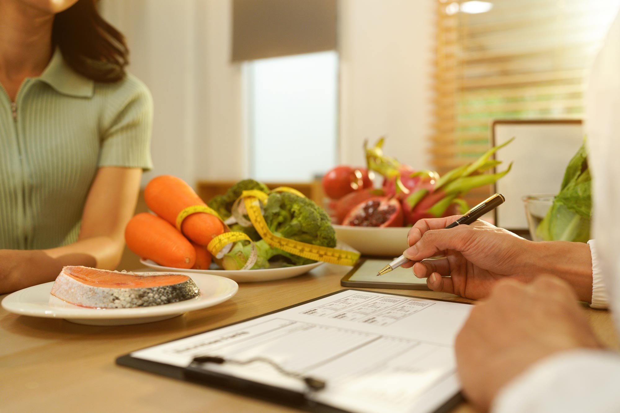 A client sits across a table from a consultant who is writing notes, with fresh vegetables, salmon and a clipboard in front of them