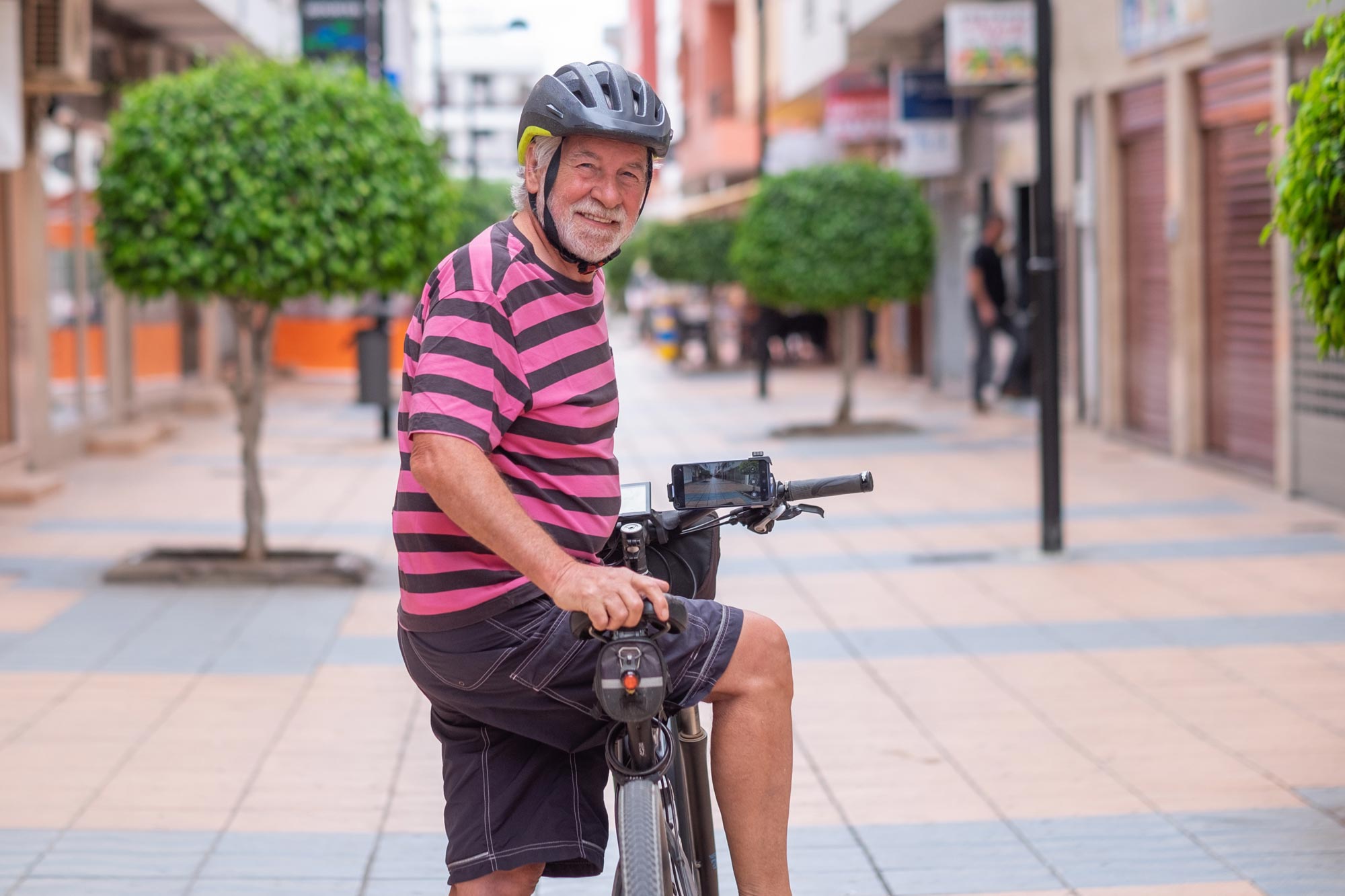 A smiling older man in a cycling helmet sits on his bike on a tree-lined pedestrian street