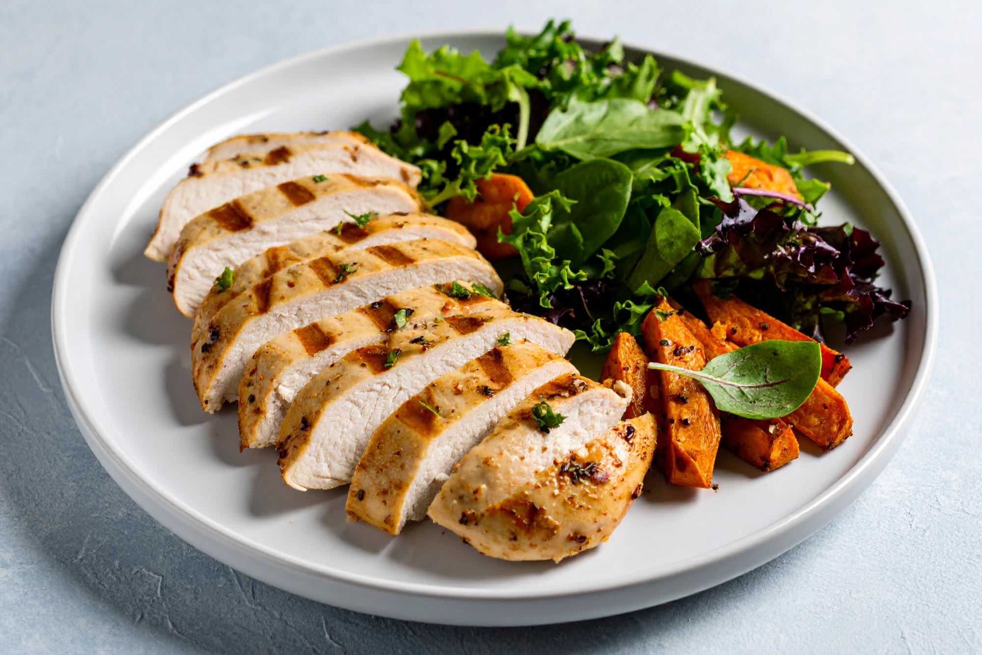 Sliced grilled chicken with salad leaves and roasted sweet potato on a white plate