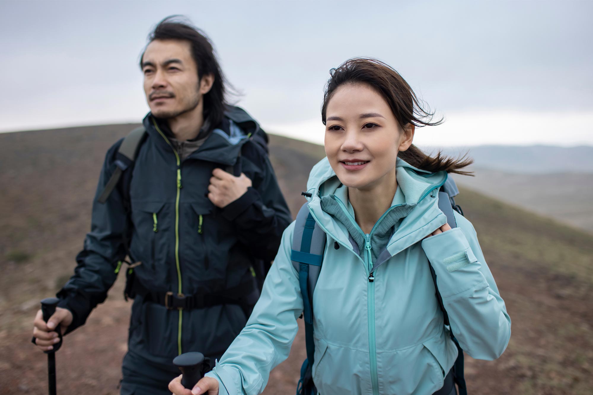 Two hikers with trekking poles walk along a hillside ridge in cool, breezy light