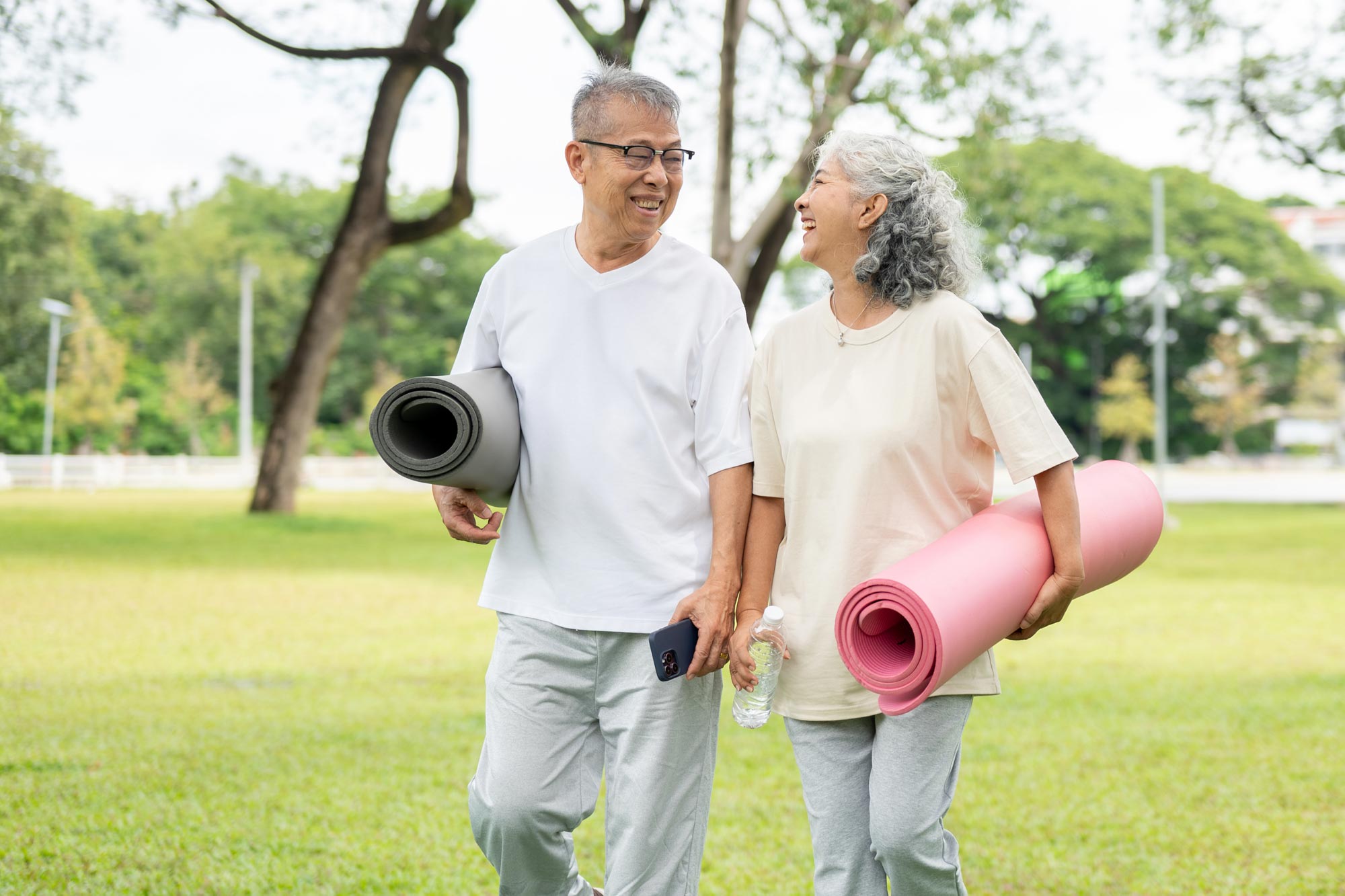 An older couple laugh together as they walk through a park carrying yoga mats and a water bottle